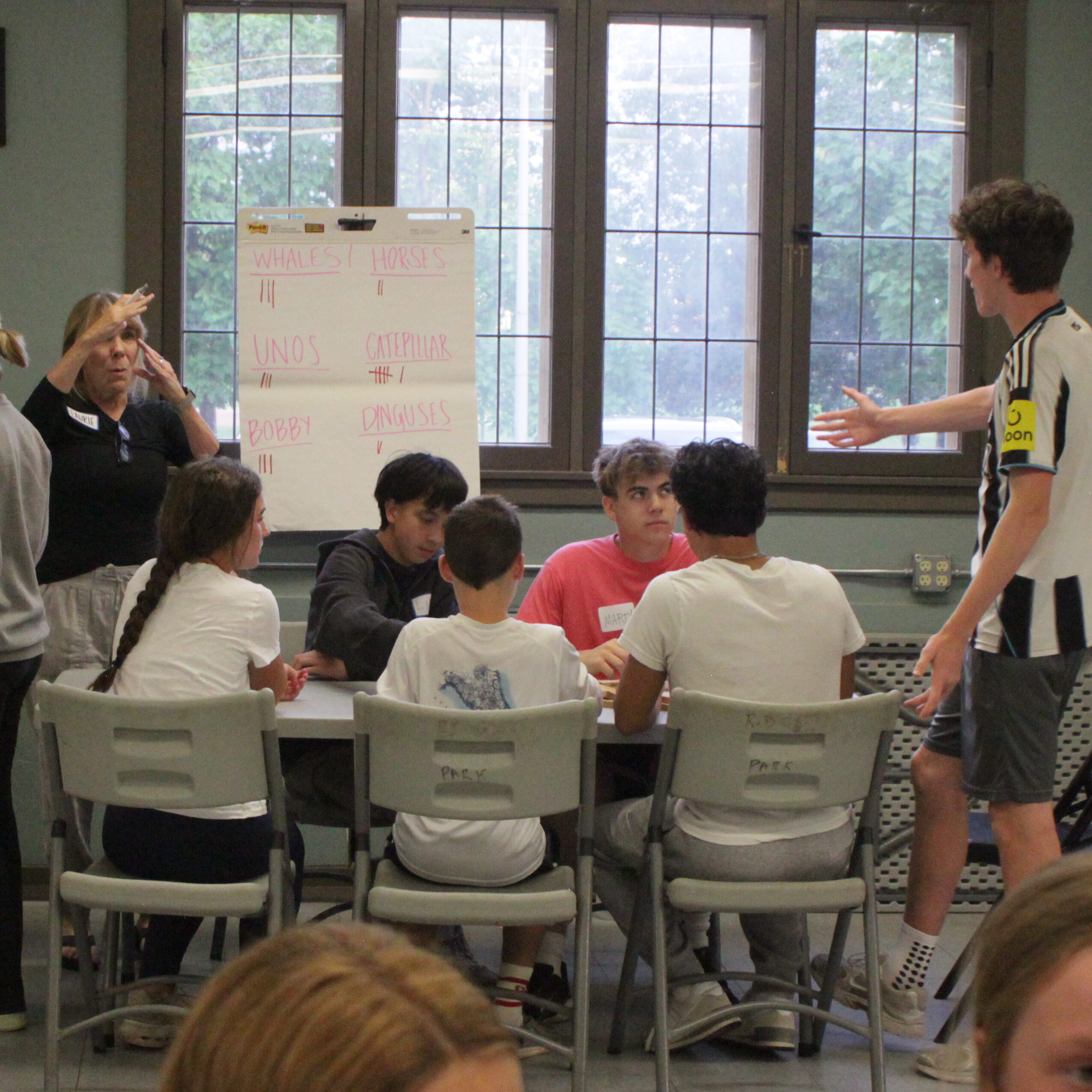 Group of youths seated around a plastic table. One youth stands gesturing to a scoreboard. A ref signals timeout with hands.