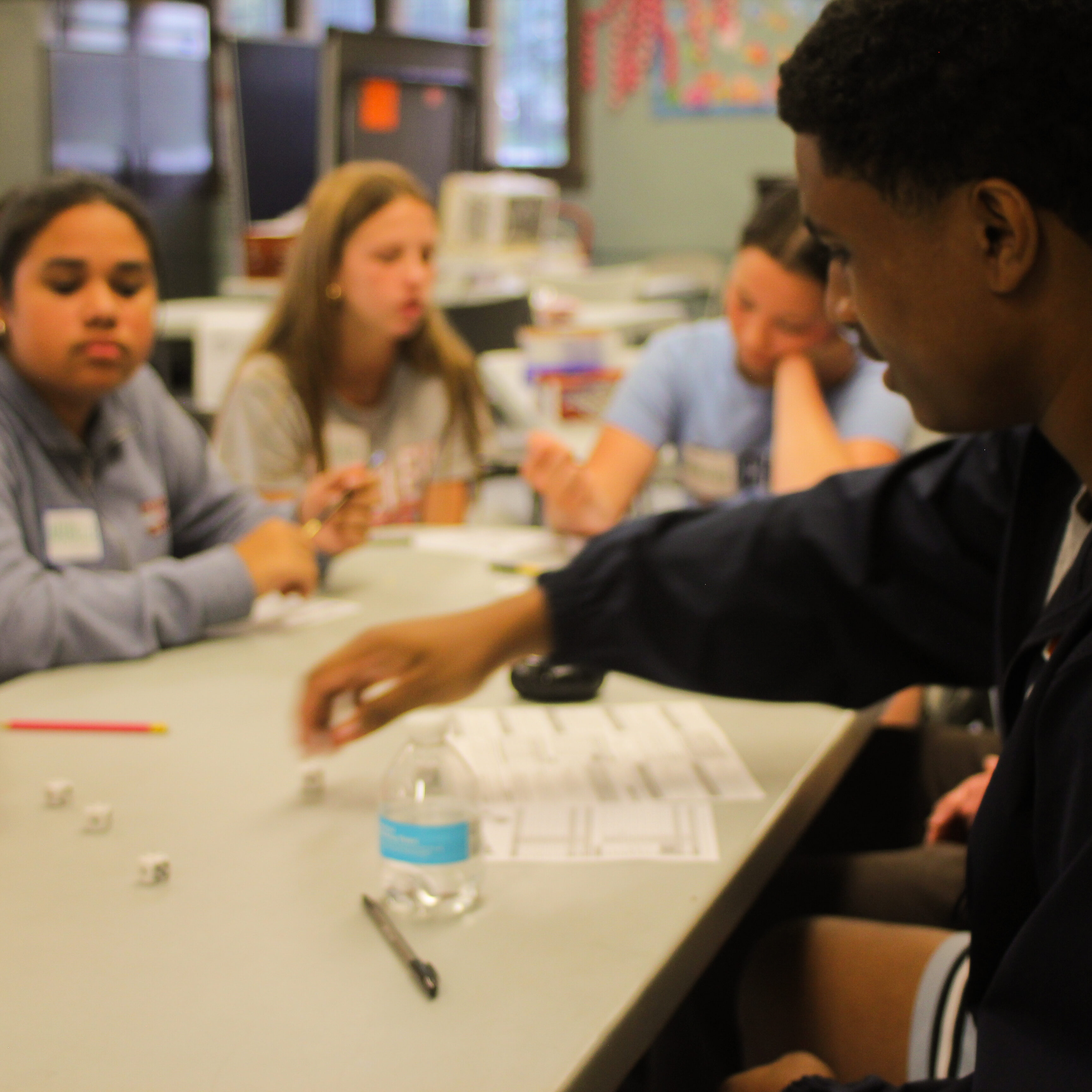 Young person rolls dice at table surrounded by peers playing Yahtzee