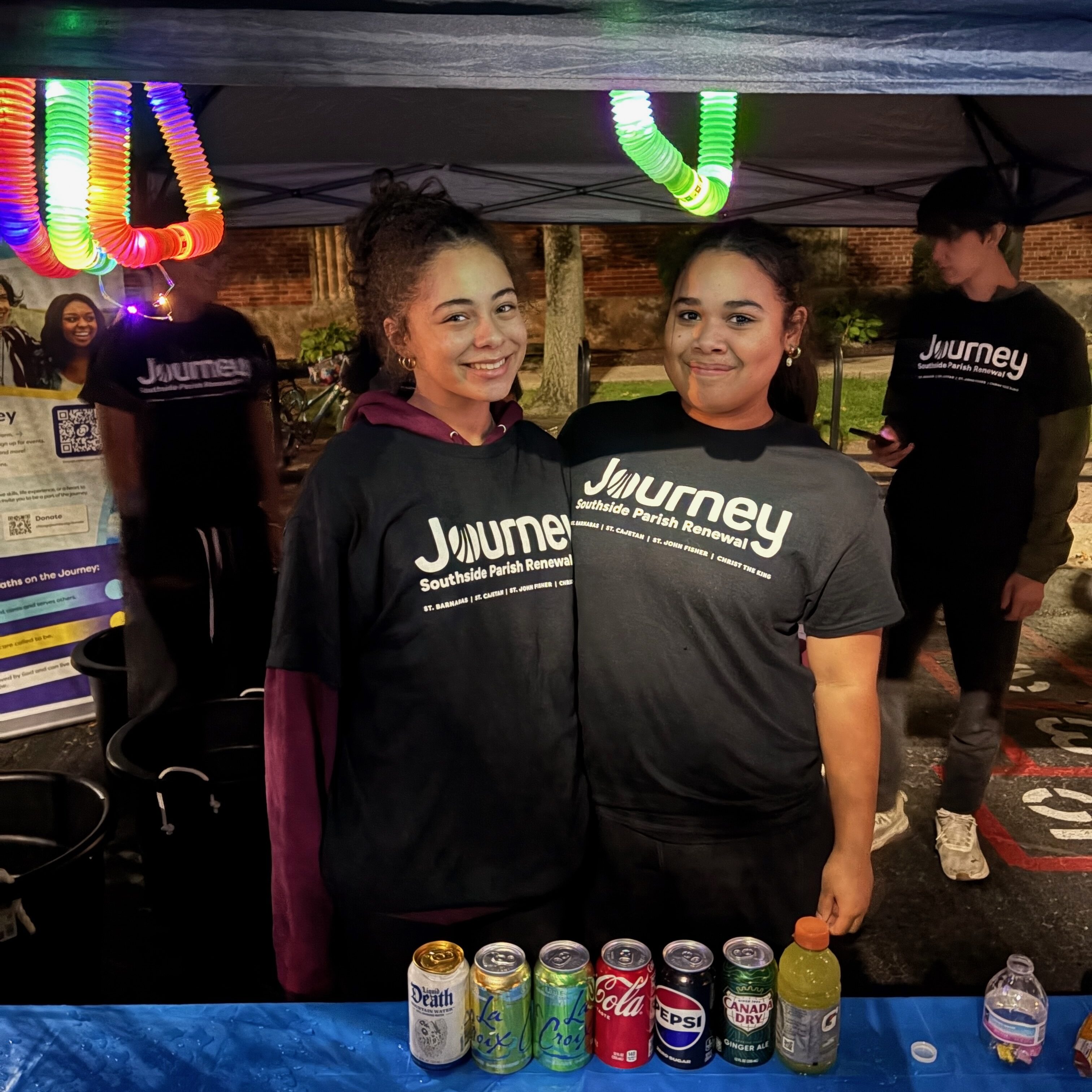 Two smiling teens in Journey t-shirts pose close together behind the drink table at night, with glowing rainbow light tubes hanging from the tent above them at St. Barnabas Fall Fest.