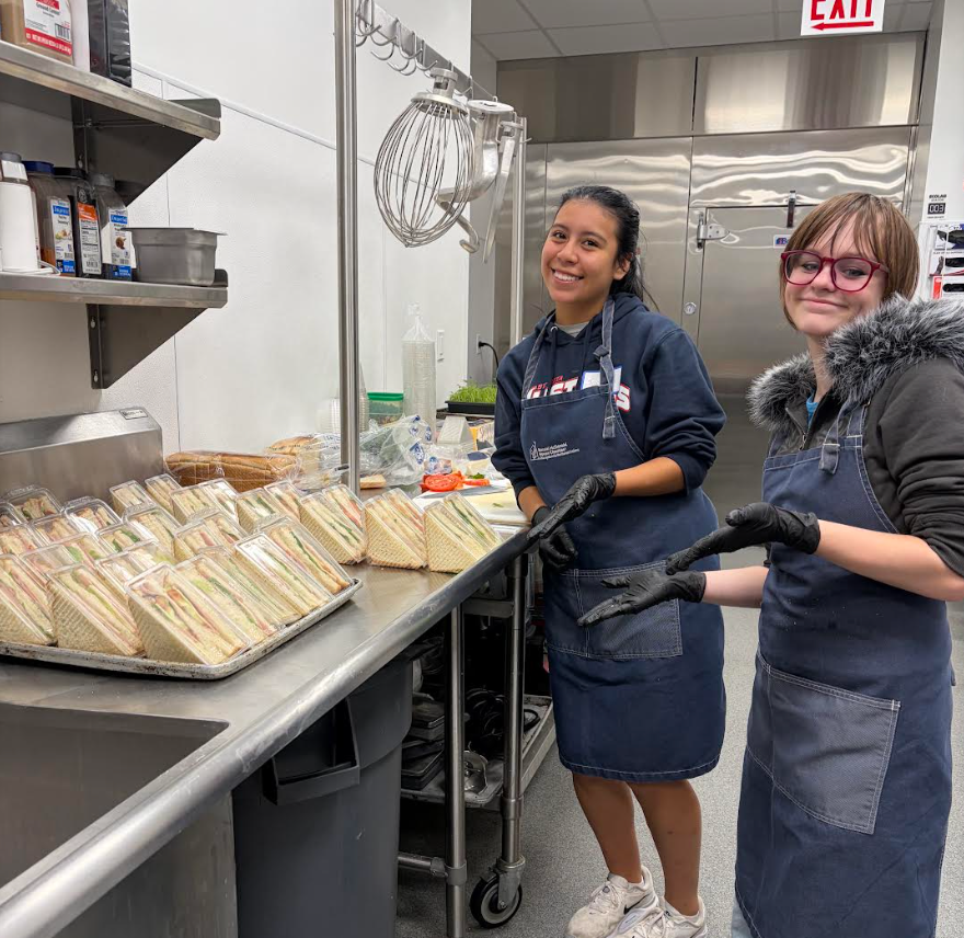 Two teen volunteers wearing gloves and aprons stand beside trays of packaged sandwiches while volunteering at Ronald McDonald House Chicago.