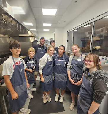 Eight teen volunteers in denim aprons pose together in a commercial kitchen during a Meals from the Heart service project at Ronald McDonald House Chicago.